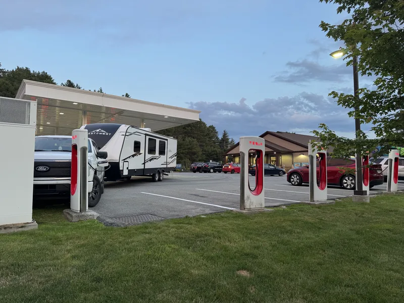 The Ford F-150 Lightning and East to West Alta at a Tesla Supercharger at dusk beside a fuel-station canopy.