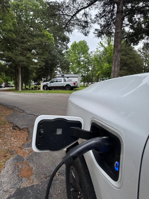 Close-up of a Ford F-150 Lightning plugged in and charging for free at a tree-shaded campsite.