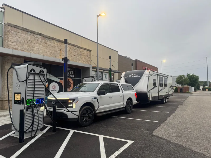 A white Ford F-150 Lightning charging from its front port at a Mercedes-Benz DC fast charger while still hitched to the 32-foot camper.