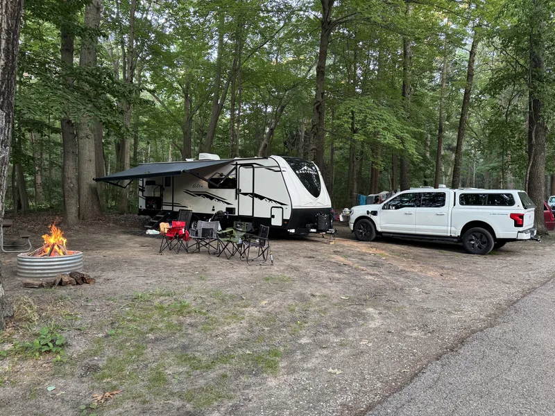 The full Muskegon State Park campsite: the East to West Alta with its awning out, camp chairs and a fire ring in front, and the white Ford F-150 Lightning parked among the trees.