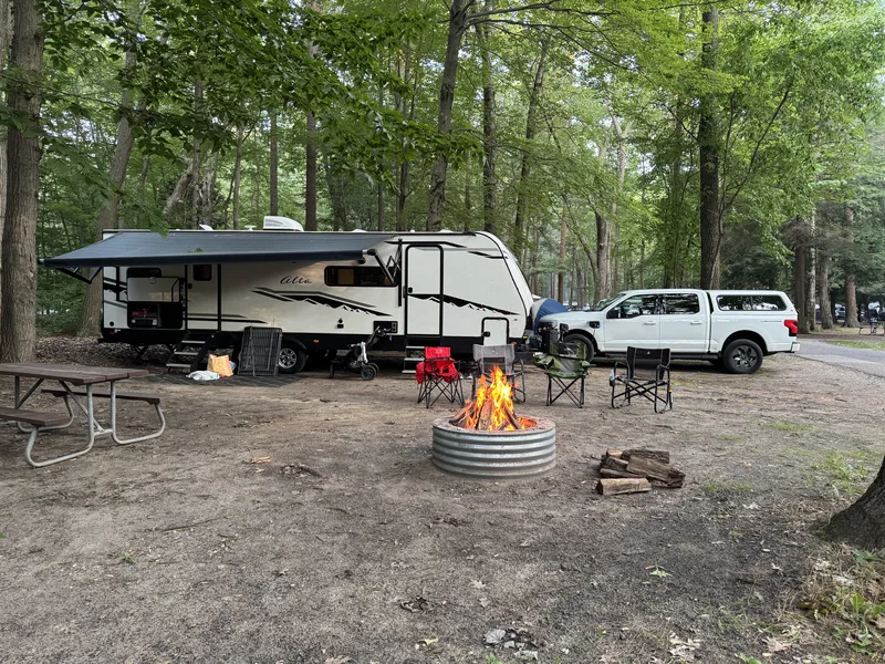 A Ford F-150 Lightning and 32-foot East to West Alta camper at a wooded Muskegon State Park campsite with a campfire going.