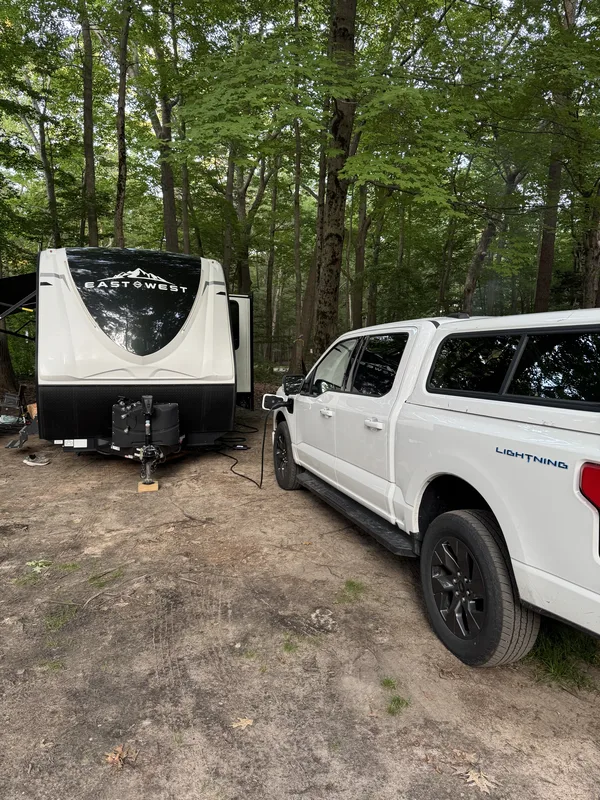 A white Ford F-150 Lightning with a bed topper parked beside a 32-foot East to West Alta travel trailer at a wooded Muskegon State Park campsite, a charging cable running to the truck.