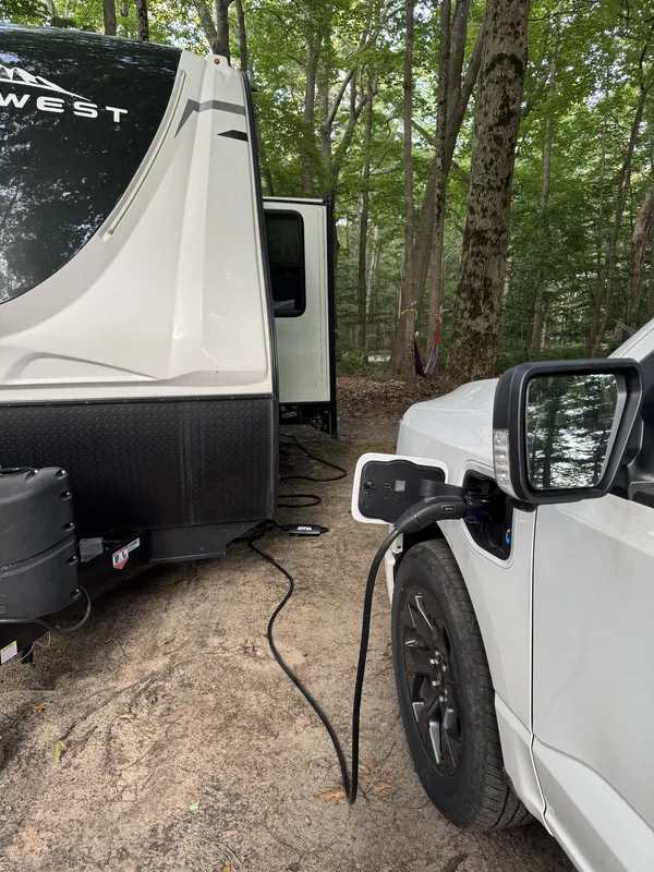 Close-up of the Ford F-150 Lightning charge port with a Jowua Mobile Connector plugged in at the wooded campsite, the East to West Alta trailer alongside.