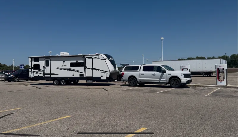 The white Ford F-150 Lightning plugged into a Tesla Supercharger with the 32-foot East to West Alta trailer still attached behind it, blue sky overhead.