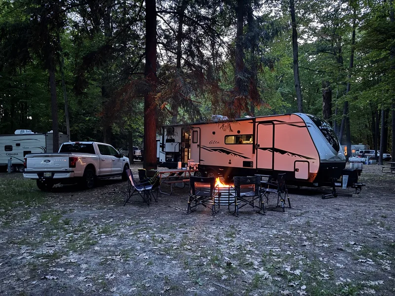 A Ford F-150 Lightning and travel trailer at a wooded campsite at dusk, a campfire glowing in front of camp chairs set up around it.