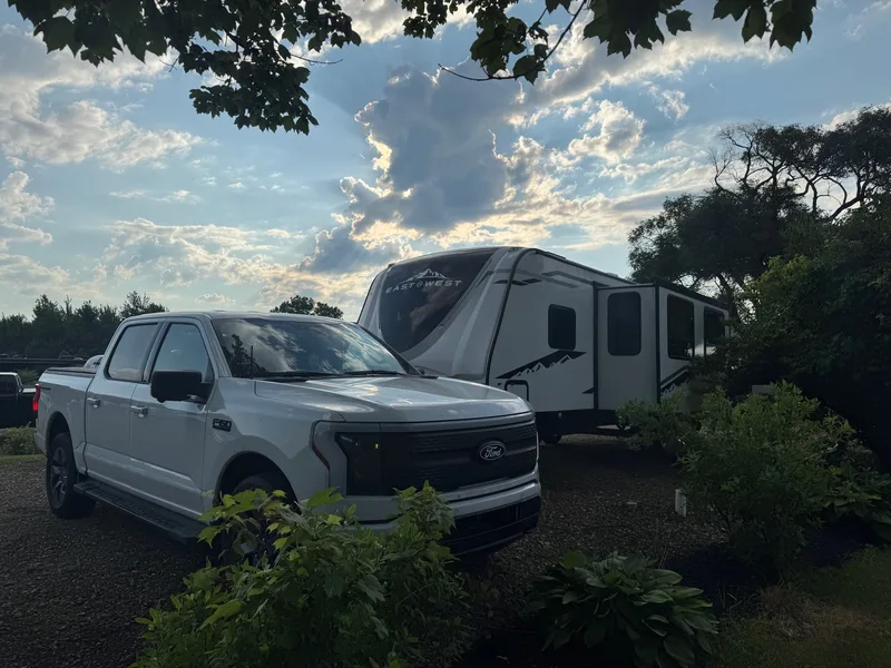 A white Ford F-150 Lightning hitched to a 32-foot East to West travel trailer at a wooded state-park campsite under a golden-hour sky.