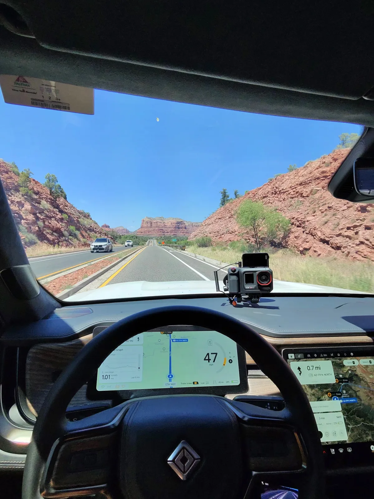 Windshield-pov shot inside a red sandstone canyon on AZ-179 north of Phoenix, dual-lane highway flanked by towering rust-red rock walls, Bell Rock visible in the distance, a GoPro mounted on the dash and the Rivian center display showing 47 mph and a 1.01 hr ETA