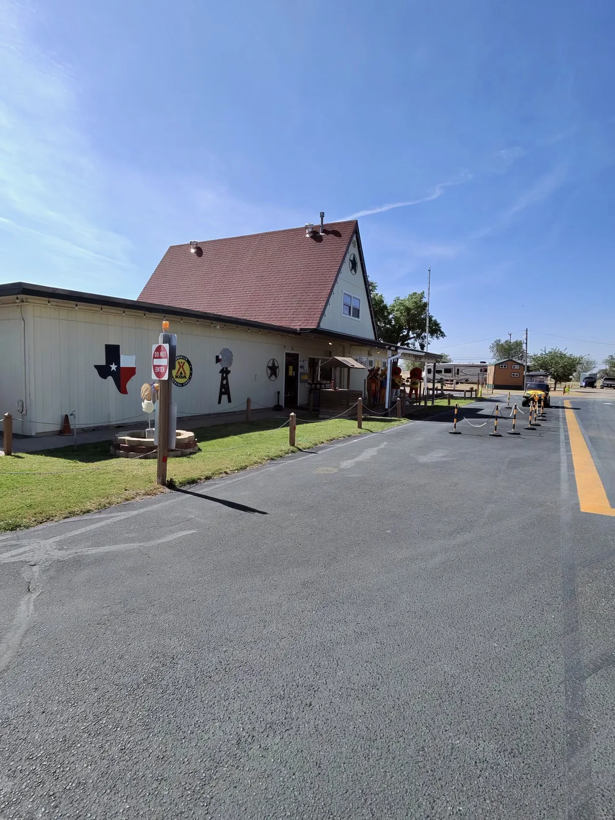 Exterior view of the Amarillo KOA Holiday office, a white-sided A-frame building with a dark red shingled roof, Texas-themed mural on the side wall featuring a Texas flag silhouette and KOA logo