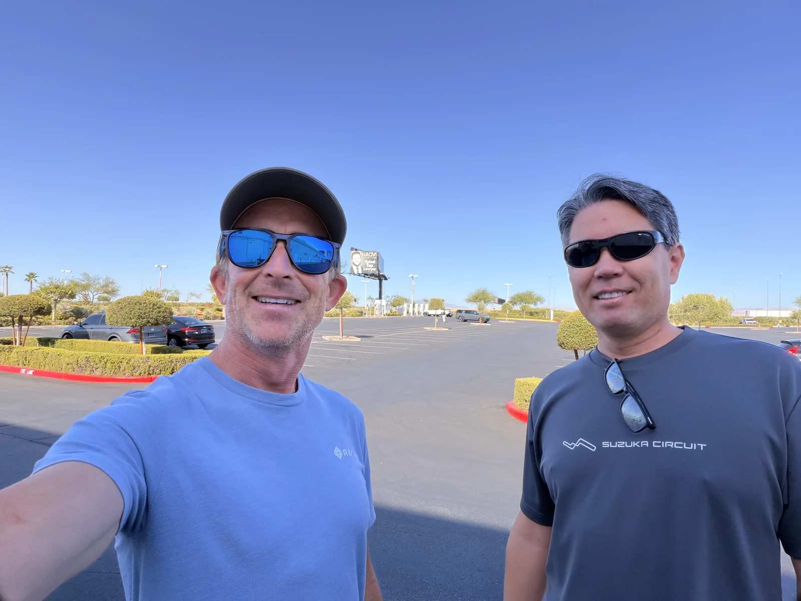 Two grinning travelers in sunglasses take a selfie at the Las Vegas Rivian charger on a bright morning, one in a Suzuka Circuit shirt, the charging plaza and palms behind.